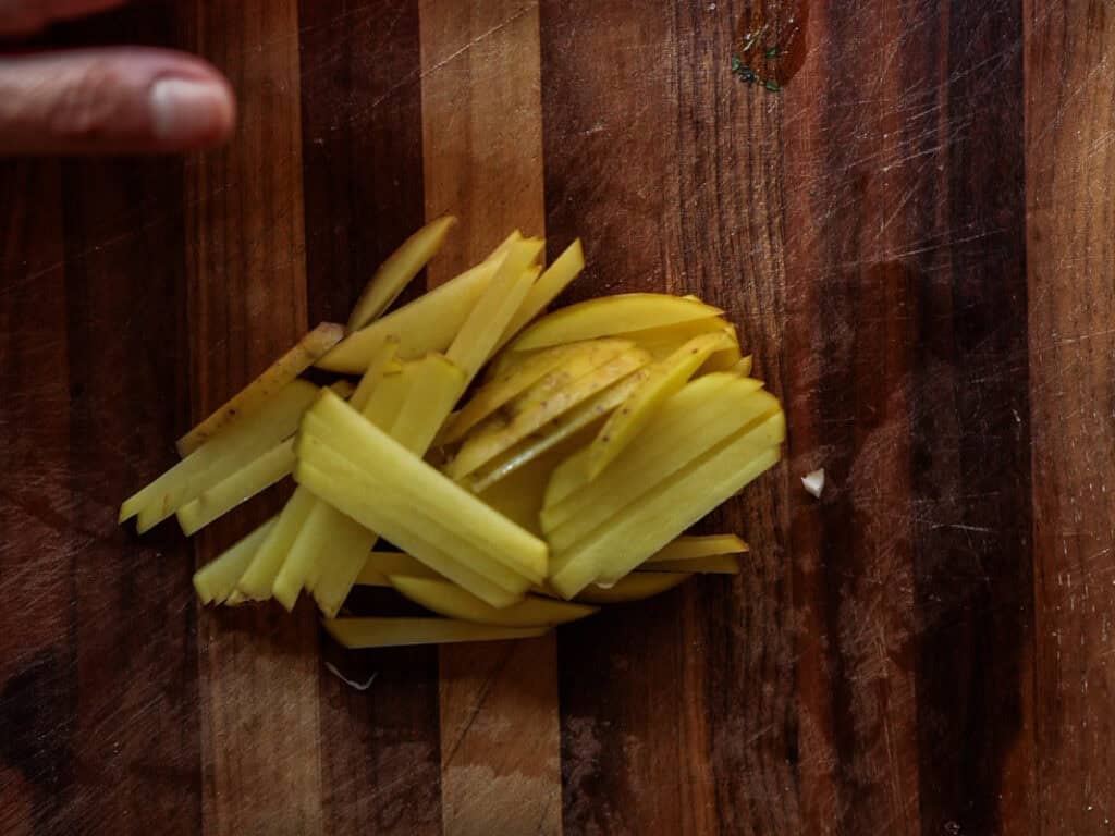 Thinly sliced potatoes are neatly arranged on a wooden cutting board. A hand is partially visible in the upper left corner, suggesting recent slicing activity. The board has a rich, dark grain pattern.